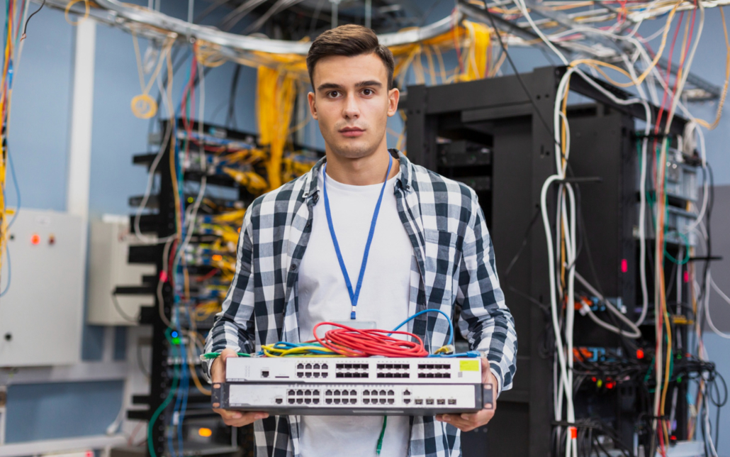 A network technician holding a server rack component in a data center, surrounded by numerous cables and networking equipment.