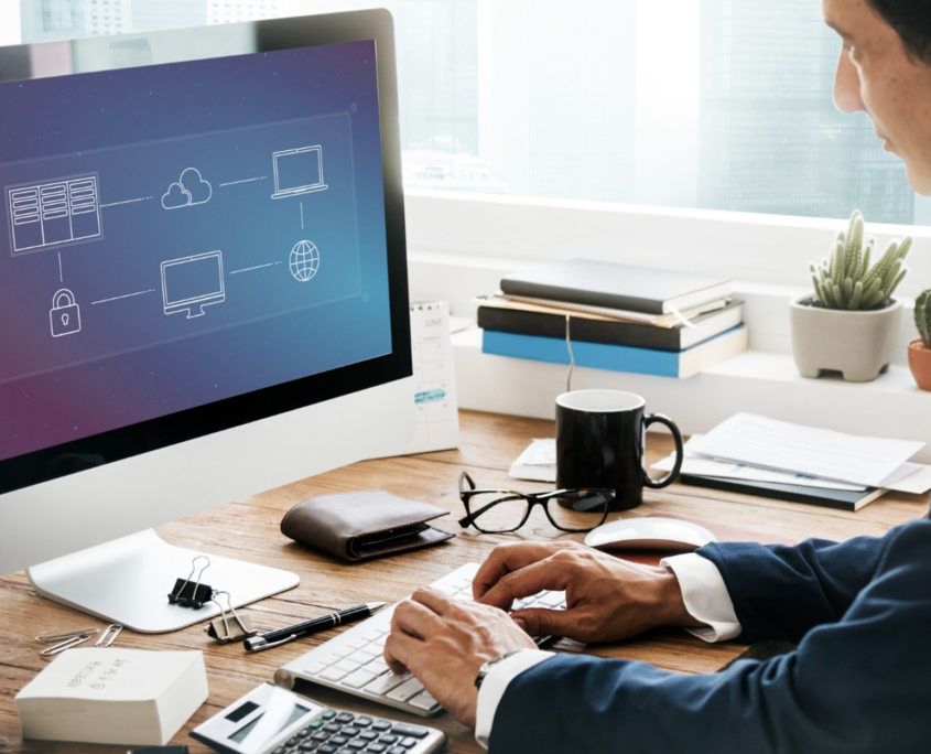 A man in a suit working at a desk with a computer displaying a network diagram, surrounded by office supplies and small potted plants.