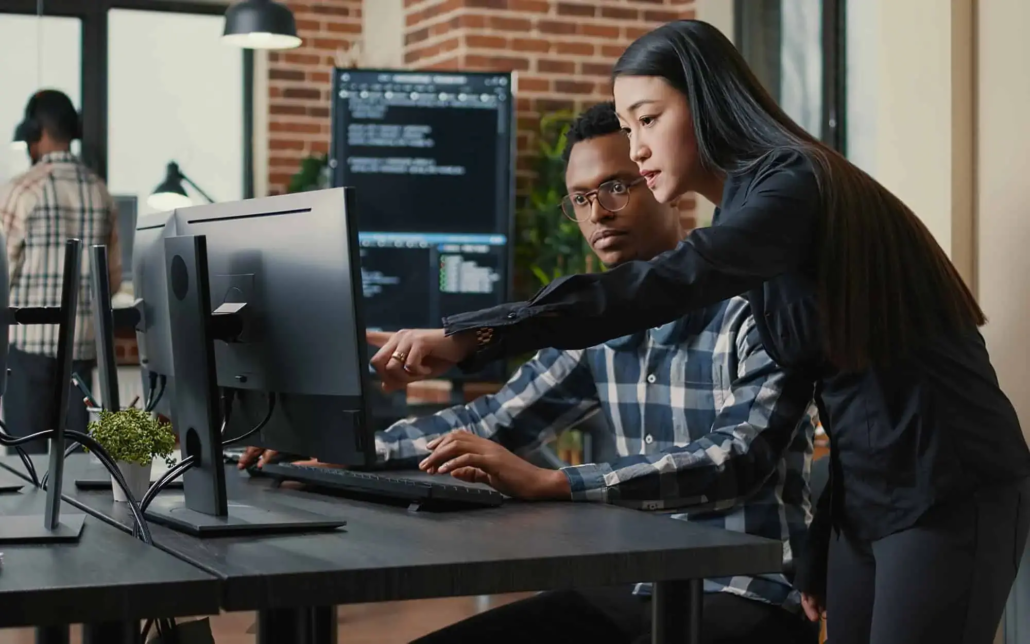 In a modern office, two colleagues collaborate on a computer. One person points at the screen while explaining something, and the other listens intently, reflecting a detailed discussion. The background features other team members working, contributing to a focused and productive work environment.