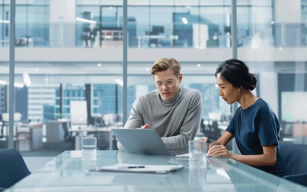 Two professionals in a conference room are intently focused on a laptop screen, sharing their thoughts and insights. They lean in close, engaged in a thoughtful discussion, with their expressions reflecting concentration and collaboration. The modern conference room setting supports their interaction, with the laptop serving as a central tool for their exchange of ideas.