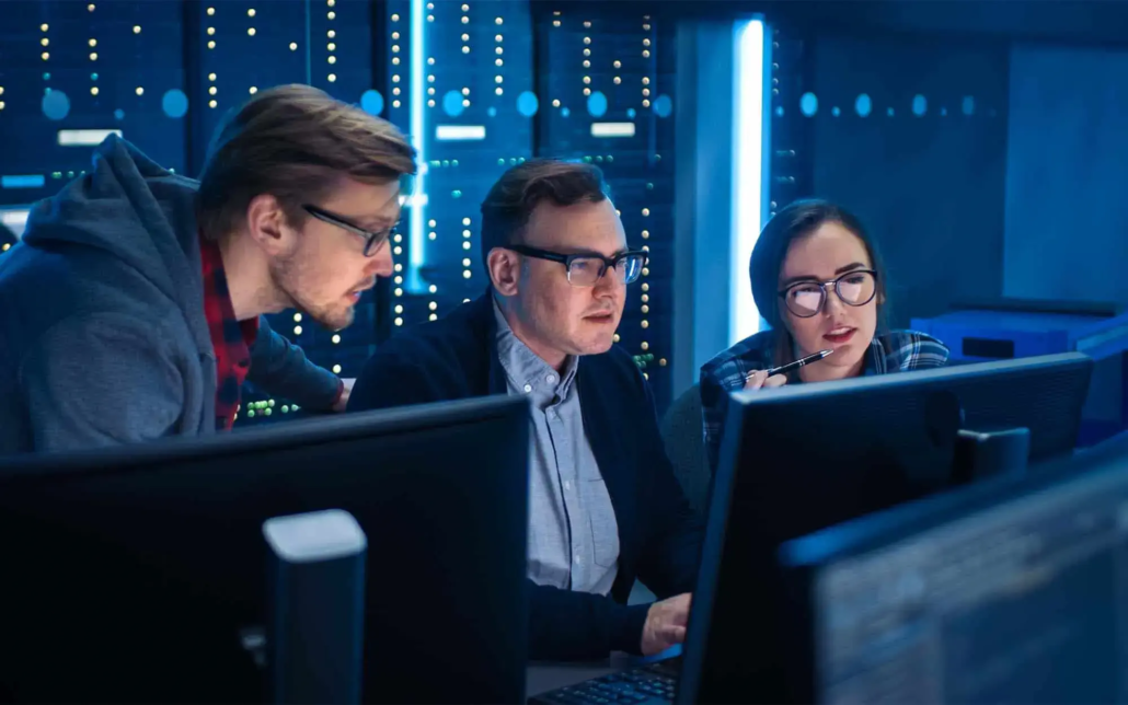 Three IT professionals collaborate in a data center, closely examining the data on their monitors. Their intense focus and teamwork are evident as they analyze information and discuss strategies, surrounded by the high-tech environment of server racks and complex equipment.